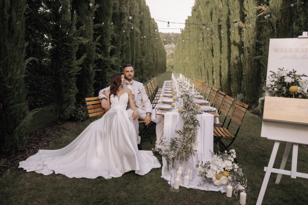Bride and groom sit together at an elegant outdoor table set for a wedding reception amidst tall trees – a perfect setting for a luxury wedding masterfully orchestrated by an experienced wedding planner.
