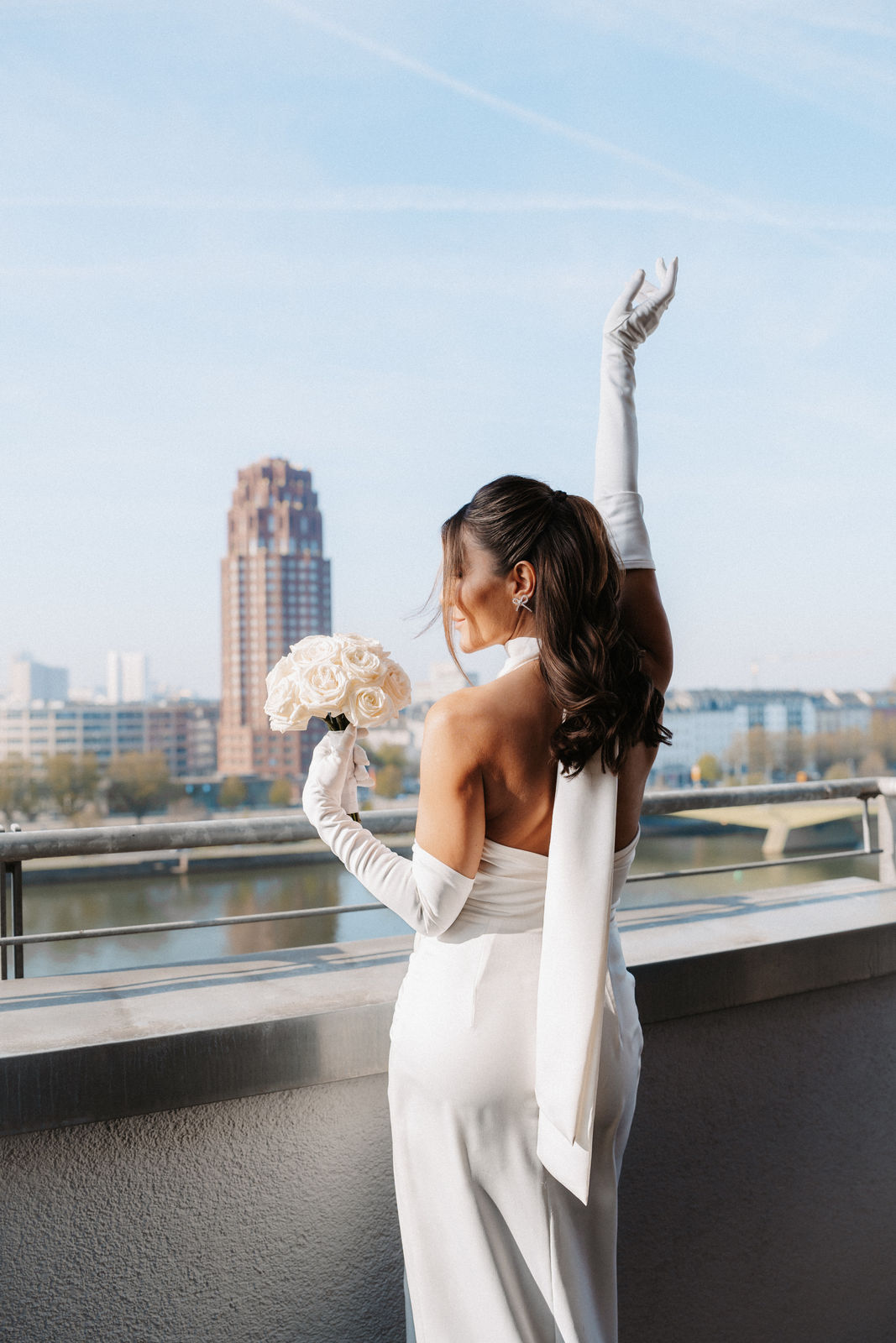 Eine Frau in einem weißen Kleid und mit Handschuhen hält einen Blumenstrauß in der Hand, hebt einen Arm und steht auf einem Balkon mit Blick auf die Stadt – ein unvergesslicher Moment, der von einer professionellen Hochzeitsplanerin für eine atemberaubende Hochzeit im Ausland inszeniert wurde.