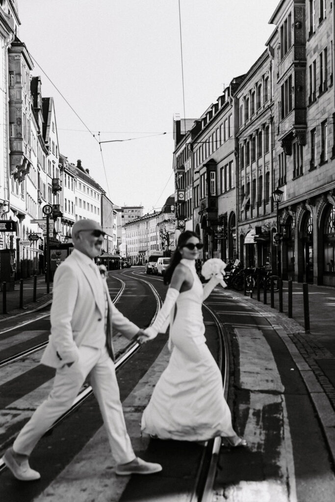 Ein Brautpaar überquert in eleganter Hochzeitskleidung Hand in Hand eine Straße in der Stadt und lächelt, während es seine Hochzeit am Reiseziel feiert.