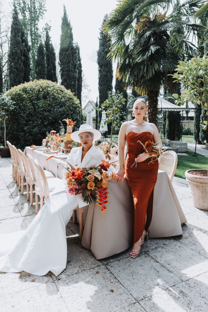 Two women in elegant dresses pose next to an open-air, festively set table with colourful floral arrangements, thereby embodying the sophistication of a „luxury wedding“ masterfully staged by a talented wedding planner.