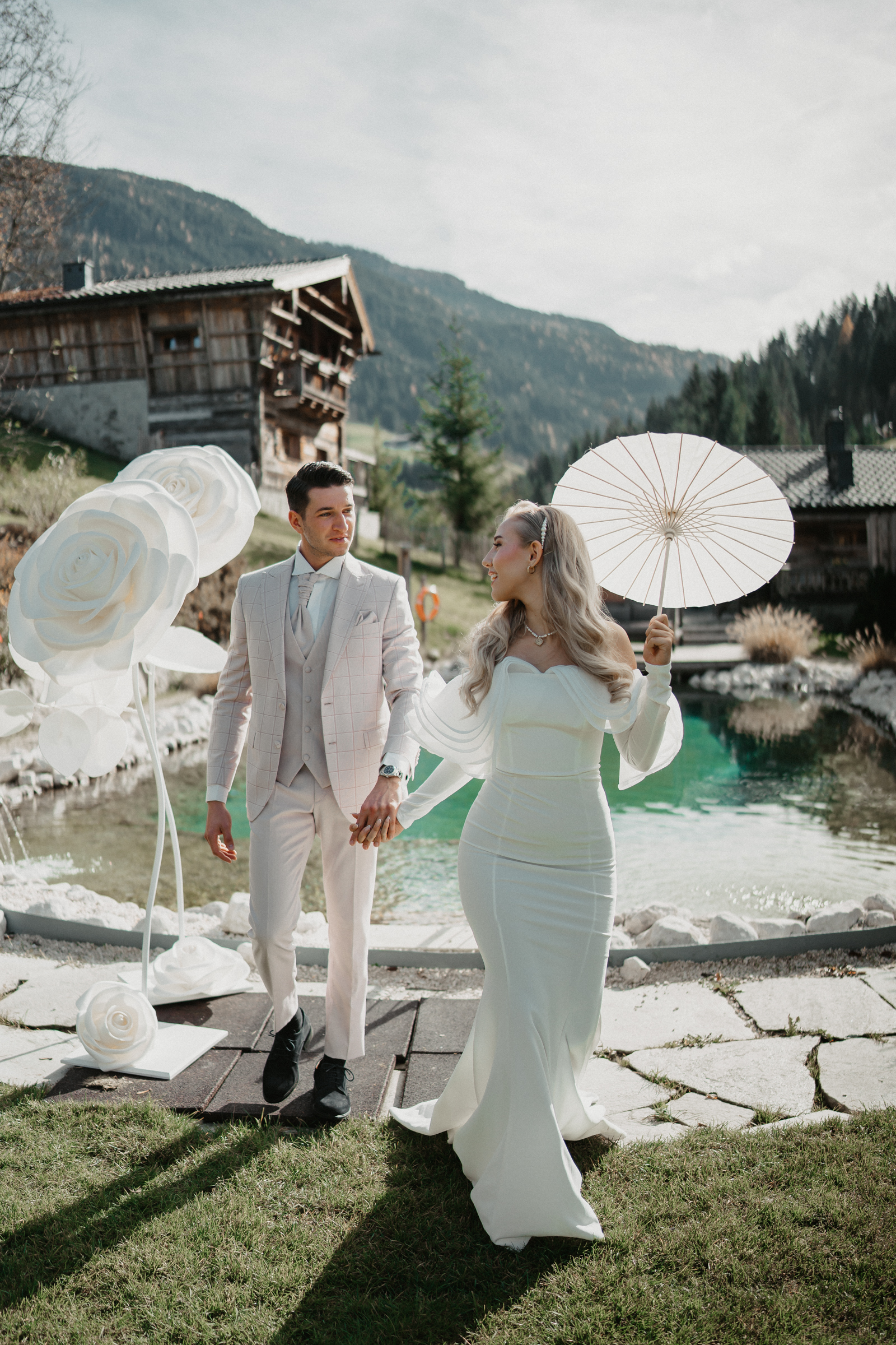 The bride in a white dress and the groom holding hands outdoors, with a white parasol and flowers on the bank of a pond, against a mountain backdrop – an unforgettable luxury wedding, perfectly organised by an experienced wedding planner, who makes every moment of this elegant event a truly magical experience.