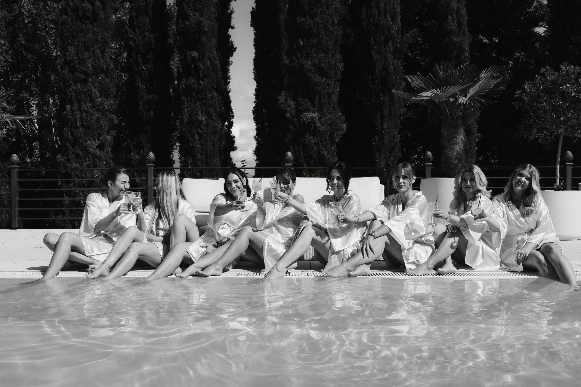 Seven women in evening dresses sit by the pool, smiling and raising their glasses, with trees visible in the background – they are celebrating a luxury wedding, organised by an experienced wedding planner, making the event a truly unforgettable experience.