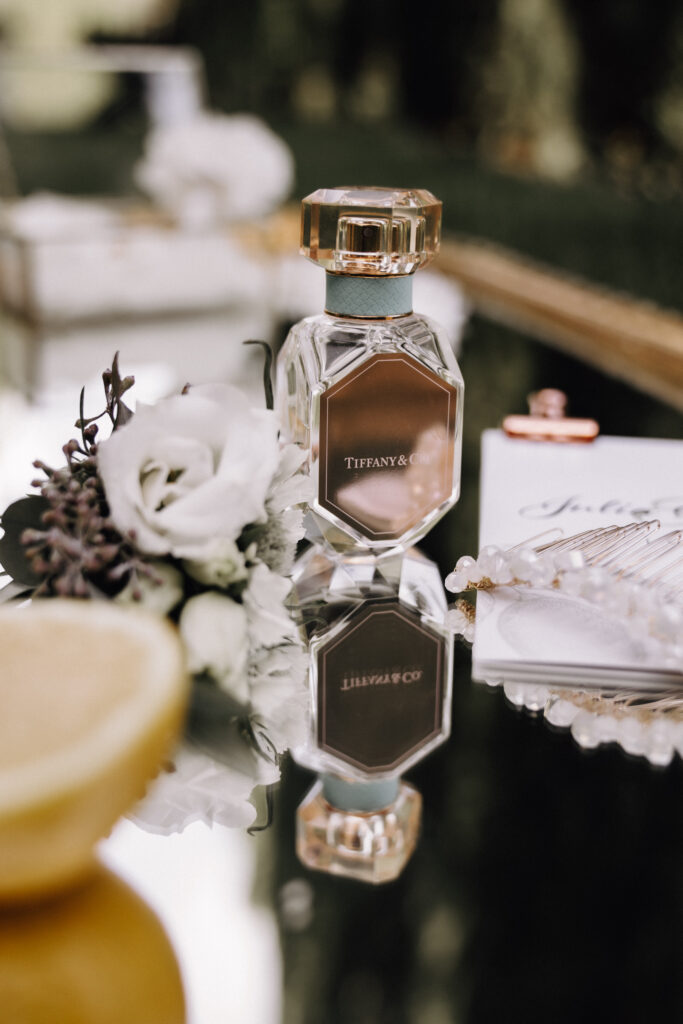 A Tiffany & Co. perfume bottle next to a white rose, a slice of lemon, pearls and an invitation on a glass surface embodies the refined elegance of a luxury wedding and captures the essence of a beautifully staged event designed by an expert wedding planner.