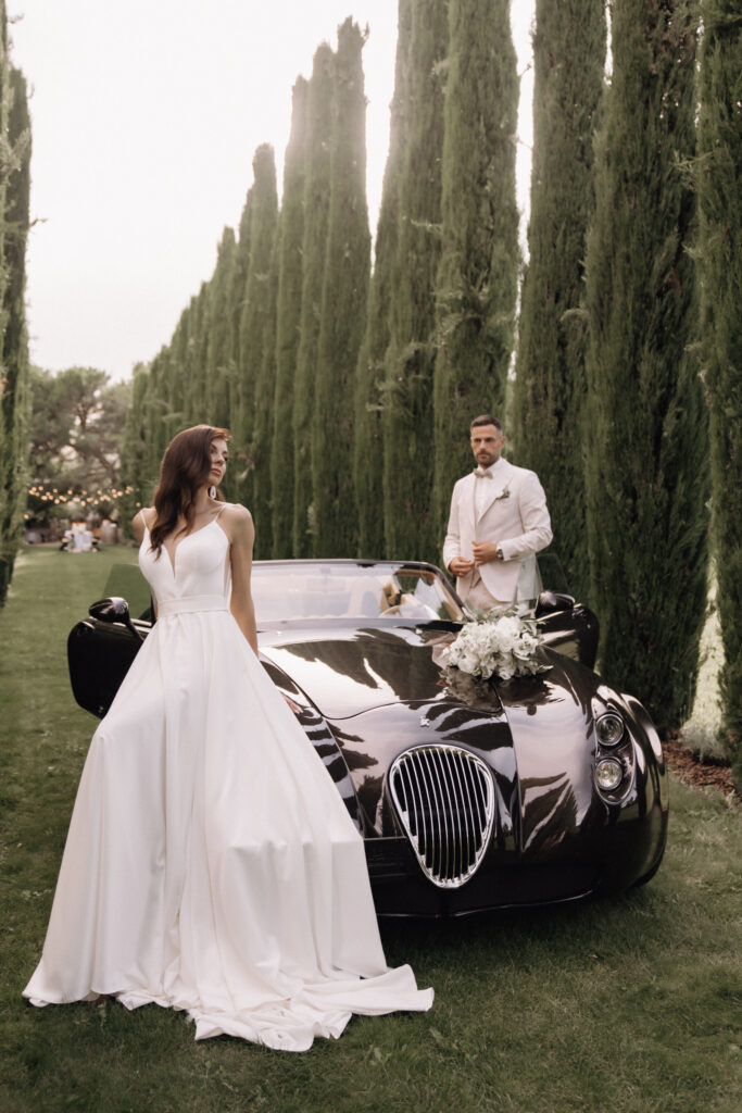 A bride in a white wedding dress stands next to a black sports car adorned with flowers; her groom in an elegant suit waits beside her – an image that captures the essence of a „luxury wedding“ and is perfect for a dream destination wedding.