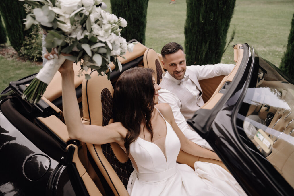 Die Braut hält ihren Blumenstrauß in der Hand, der Bräutigam fährt in einem Cabrio – beide lächeln an ihrem Hochzeitstag im Freien und feiern ihre „Luxus-Hochzeit“, die von einer erfahrenen Hochzeitsplanerin organisiert wurde.
