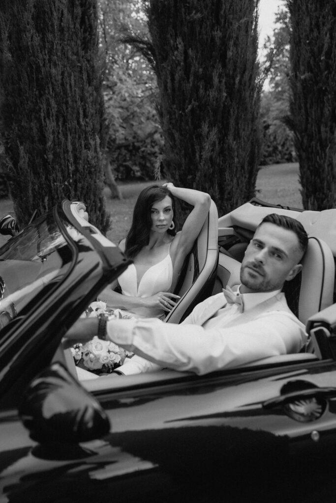 A woman and a man in festive attire sit in a convertible, looking at the camera, surrounded by trees – a scene capturing the elegance of a destination wedding.