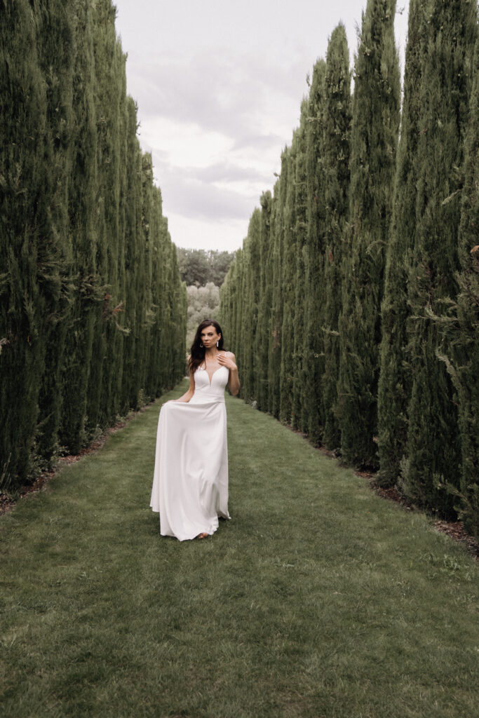 A woman in a white dress stands on a grassy path amongst tall, slender green trees beneath a cloudy sky, embodying the elegance of a wedding abroad organised by an experienced wedding planner, who turns this occasion into a truly unforgettable experience.