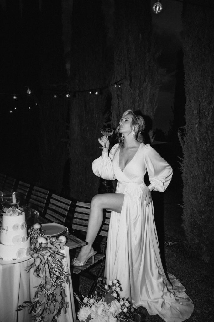 A woman in a long dress drinks from a glass while standing outdoors at night next to a festively laid table with a cake – a magical moment, staged by an experienced wedding planner, perfect for an unforgettable destination wedding.